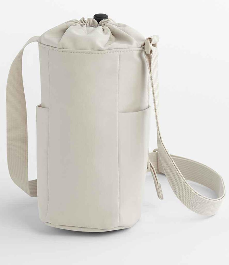 May include: Off-white, cylindrical crossbody bag with a drawstring closure and a black toggle. The bag has two small side pockets and a long, adjustable strap. The bag is made of a smooth, matte material.