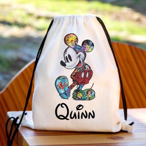 May include: A white drawstring backpack featuring a large graphic of a cartoon mouse silhouette filled with various colorful character illustrations. The name Quinn is printed in black lettering below the central character design.