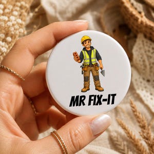 Könnte beinhalten: Ein runder weißer Button mit einer Cartoon-Illustration eines Bauarbeiters in gelbem Schutzhelm und Warnweste, der einen Ziegelstein und Werkzeuge hält. Der Text MR FIX-IT ist in fetten schwarzen Großbuchstaben unter der Figur gedruckt.