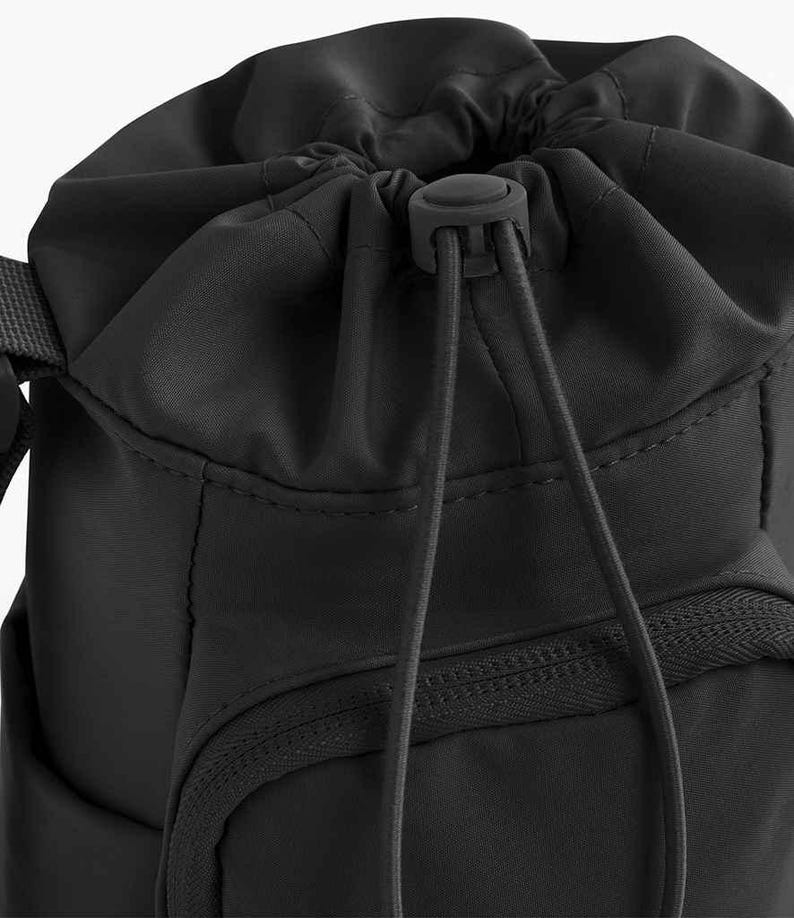 May include: Black drawstring backpack with a zippered front pocket. The bag has a rounded top with a drawstring closure and a black plastic toggle. The bag is made of a smooth, matte fabric.