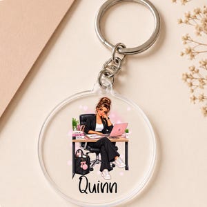 Op de afbeelding: Een ronde sleutelhanger van doorzichtig acryl met een bedrukte illustratie van een persoon met bruin haar in een zwarte blazer en broek, zittend aan een bureau met een laptop en boeken. De naam Quinn staat onderaan in zwarte letters.