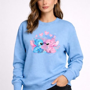 Puede incluir: Sudadera azul claro con cuello redondo y mangas largas. Presenta una ilustraci&oacute;n en acuarela de dos personajes de dibujos animados, uno azul y otro rosa, rodeados de corazones rosas.