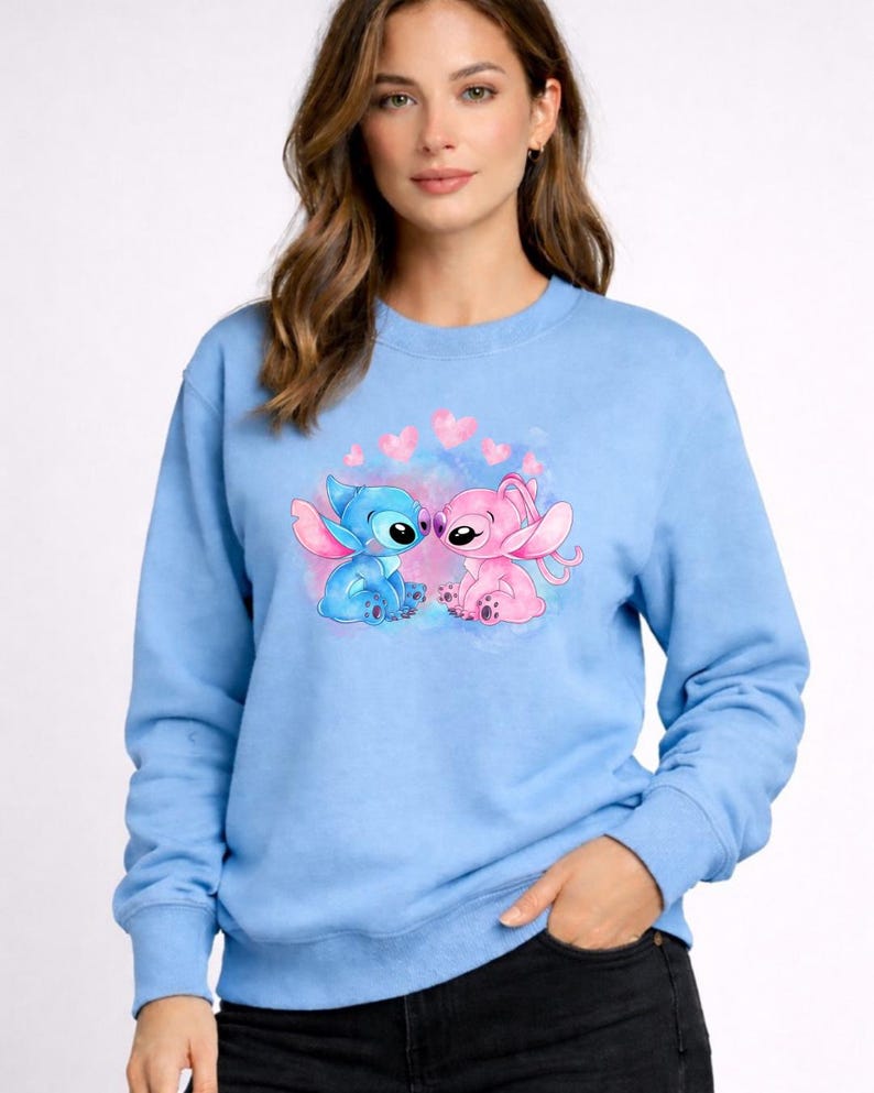 Puede incluir: Sudadera azul claro con cuello redondo y mangas largas. Presenta una ilustraci&oacute;n en acuarela de dos personajes de dibujos animados, uno azul y otro rosa, rodeados de corazones rosas.