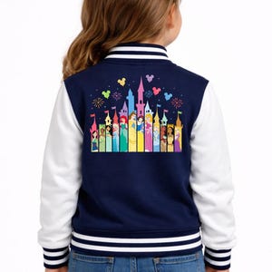 May include: Navy and white varsity jacket with a colourful graphic of princesses and castles. The jacket has white sleeves, a striped collar, and a striped hem. The graphic includes fireworks and heart-shaped designs.