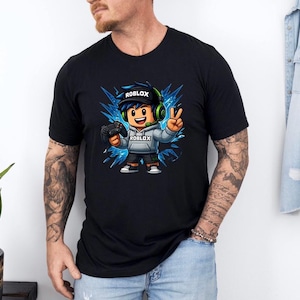 May include: Black t-shirt featuring a cartoon character with the Roblox logo on the hat and hoodie. The character is holding a game controller and making a peace sign. The design includes blue and white accents, and the word "ROBLOX" is visible.