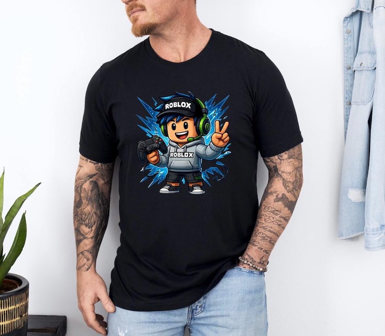 May include: Black t-shirt featuring a cartoon character with the Roblox logo on the hat and hoodie. The character is holding a game controller and making a peace sign. The design includes blue and white accents, and the word "ROBLOX" is visible.