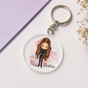 Peut inclure: Un porte-clés circulaire en acrylique transparent présentant une illustration de personnage aux longs cheveux bruns, vêtu d'un haut noir et d'un pantalon noir. Le motif inclut des touches d'aquarelle rose, des détails floraux et le nom Quinn en noir.