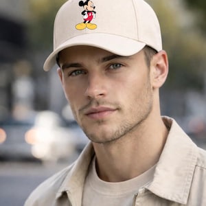 May include: A beige baseball cap with a Mickey Mouse graphic. The cap has a curved peak and a structured design. The Mickey Mouse illustration shows the character in his iconic red shorts, yellow shoes, and black gloves.