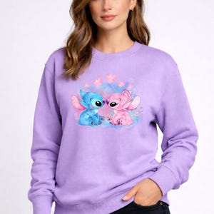 Puede incluir: Una sudadera lavanda con un dise&ntilde;o de los personajes Stitch y Angel de la pel&iacute;cula de Disney Lilo & Stitch. Stitch azul y Angel rosa se representan abrazados, rodeados de corazones rosas.