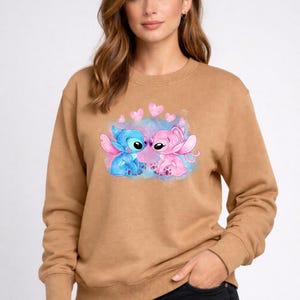 Puede incluir: Una sudadera marr&oacute;n con un gr&aacute;fico de dos personajes de dibujos animados, uno azul y otro rosa, con corazones encima. Los personajes est&aacute;n sobre un fondo de acuarela azul y rosa. La sudadera tiene mangas largas.