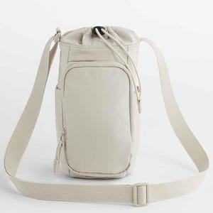May include: A beige crossbody bag with a drawstring closure at the top. The bag has a front pocket with a zipper and an adjustable shoulder strap. The bag is made of a smooth, durable material.