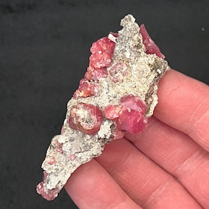 May include: A natural mineral specimen featuring vibrant, deep pink crystals clustered on a matrix of grey and beige rock. The crystals have a glossy appearance, contrasting with the rough texture of the host rock. The specimen is held against a dark background.