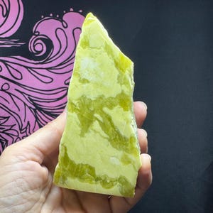 Può includere: Una mano tiene una grande pietra di serpentino grezza, verde chiaro, con venature verde scuro. La pietra ha un bordo naturale ruvido e una superficie lucida. Lo sfondo è nero con un motivo floreale rosa e bianco.