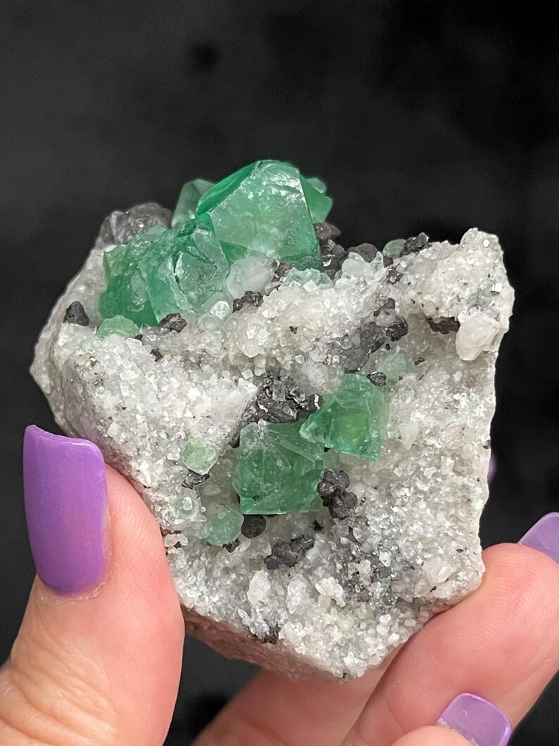 K&ouml;nnte beinhalten: Ein Cluster gr&uuml;ner Fluoritkristalle auf einer wei&szlig;en Matrix mit schwarzen Einschl&uuml;ssen. Die Fluoritkristalle sind scharf und gut definiert, und die Matrix ist rau und strukturiert.