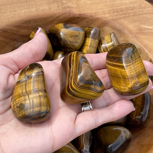May include: A collection of tiger's eye tumbled stones in various shades of brown and gold. The stones have a smooth, polished surface and are arranged in a bowl.
