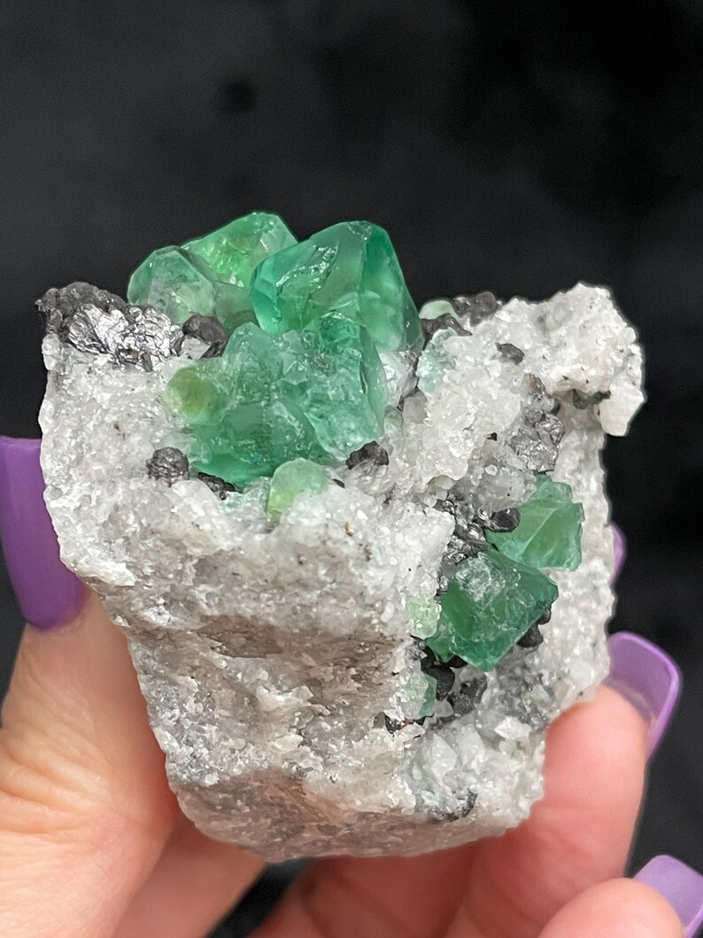 K&ouml;nnte beinhalten: Ein Cluster gr&uuml;ner Fluoritkristalle auf einer Matrix aus wei&szlig;em Calcit und grauem Bleiglanz. Die Fluoritkristalle sind scharf und gut ausgebildet, mit einem glasartigen Glanz.