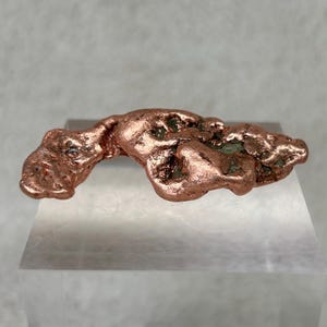 May include: A raw copper specimen with a textured, organic shape. The copper has a rich, reddish-brown color with hints of green oxidation. The specimen is displayed on a clear, angled surface, showcasing its natural form.
