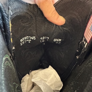 May include: Black leather cowboy boot interior with size and style information. The boot has a stitched design and a small American flag tag. The size is 8.5M.