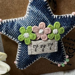May include: A denim star-shaped keyring with white stitching and a cream strap. The star is decorated with embroidered green and pink flowers and a small rectangular tag with the number "777". Pink and green beads are attached to the edges.