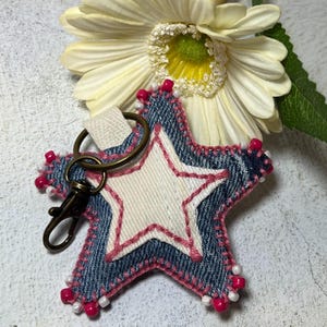 May include: A denim star-shaped keychain with a white fabric centre and pink stitching. The star is decorated with white and pink beads.