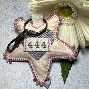May include: A white star-shaped fabric keyring with pink trim and beads. The keyring has a heart-shaped patch with a plaid pattern and a tag that says "444".
