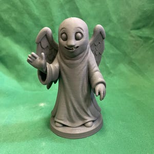 May include: A gray 3D-printed figurine of a smiling angel with wings and a flowing robe. The angel is standing on a circular base, with one hand raised in a welcoming gesture. The background is a solid green color.