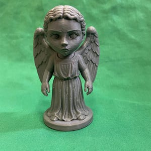 Puede incluir: Figura gris de un ángel con grandes alas, una túnica fluida y una expresión solemne. La estatua está sobre una base circular. El cabello del ángel está peinado en ondas.