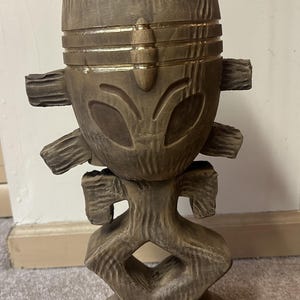 May include: A brown wooden sculpture with a carved design. The sculpture has a stylized face with large eye shapes and gold accents. It has a textured surface and outstretched arms. The base is round.