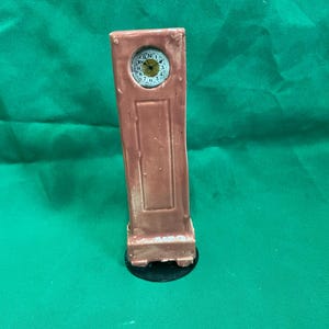 Könnte beinhalten: Eine kleine, braune Standuhr mit weißem Zifferblatt und schwarzen Zeigern. Die Uhr hat einen rechteckigen Körper und steht auf einem kleinen schwarzen Sockel. Das Zifferblatt zeigt die Zahlen 1 bis 12.