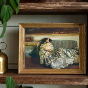 May include: A framed painting of a woman in a white dress lying on a couch. The painting is in a gold frame and is hanging on a wooden shelf.