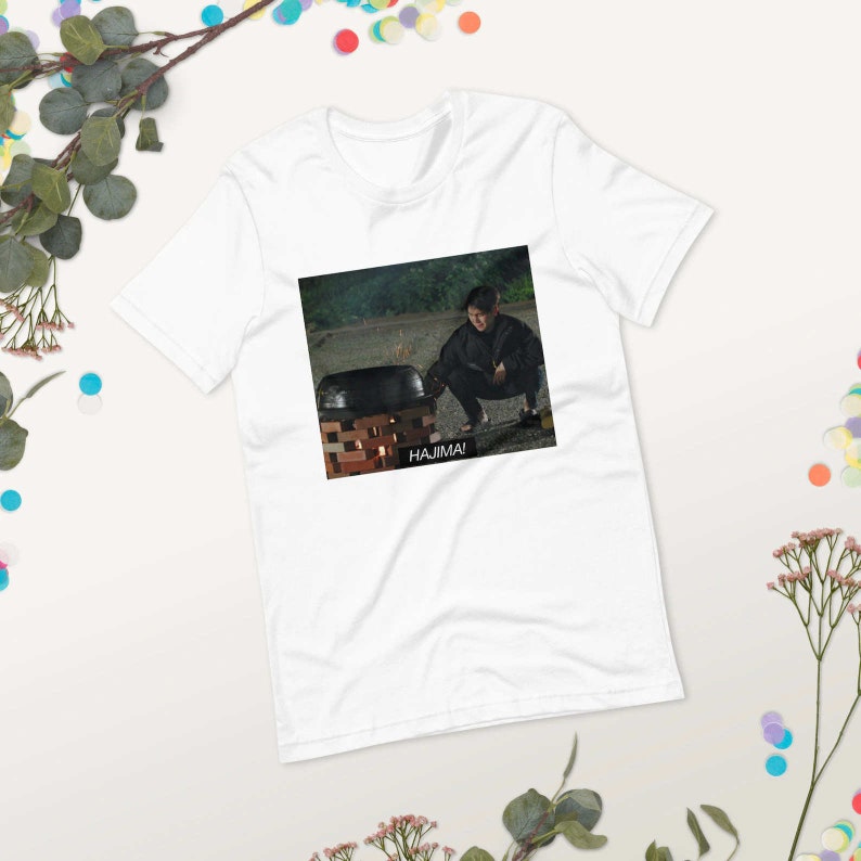 May include: White t-shirt with a black and white image of a person in a black jacket and black pants squatting in front of a black pot on a brick base with flames underneath. The text "HAJIMA!" is printed below the image.