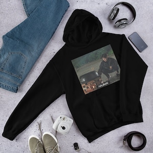 May include: Black hooded sweatshirt with a graphic print of a person in a dark green jacket and black pants kneeling in front of a pot with the text "HAJIMA!" printed below the image.