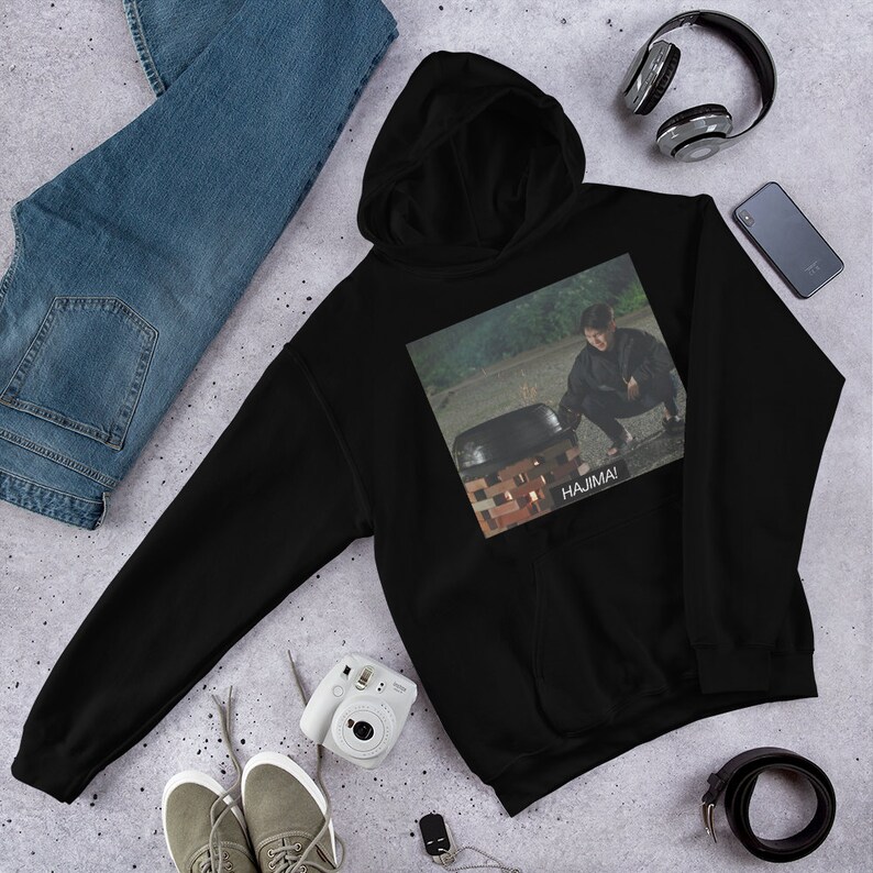 May include: Black hooded sweatshirt with a graphic print of a person in a dark green jacket and black pants kneeling in front of a pot with the text "HAJIMA!" printed below the image.