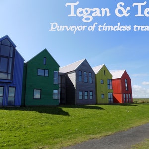 Może przedstawiać: Rząd kolorowych dom&oacute;w z niebieskim niebem i zieloną trawą na pierwszym planie. Tekst "Tegan & Teak Purveyor of timeless treasures" znajduje się w g&oacute;rnej części obrazu.