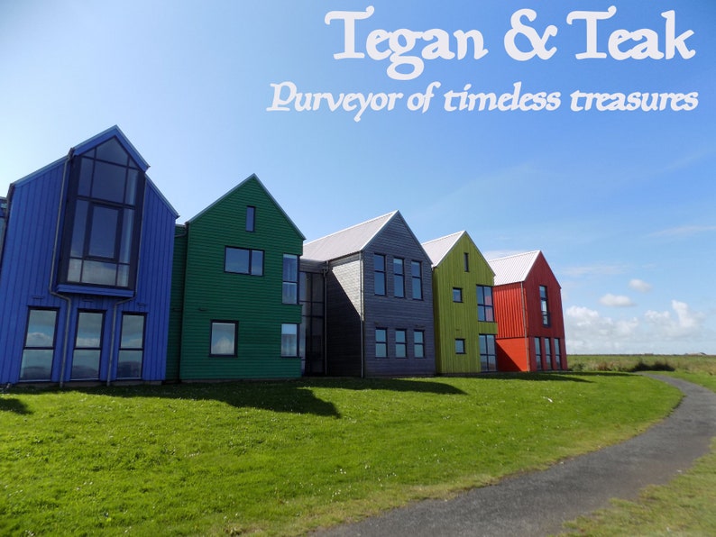 Może przedstawiać: Rząd kolorowych dom&oacute;w z niebieskim niebem i zieloną trawą na pierwszym planie. Tekst "Tegan & Teak Purveyor of timeless treasures" znajduje się w g&oacute;rnej części obrazu.