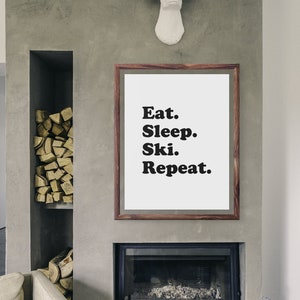 Op de afbeelding: Een ingelijst print met de tekst "Eat. Sleep. Ski. Repeat." hangt aan een grijze betonnen muur boven een open haard. De open haard heeft een zwarte metalen omlijsting en een zwarte glazen deur. Boven de print is een wit hertenhoofd aan de muur bevestigd.