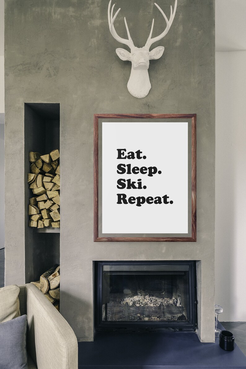 Op de afbeelding: Een ingelijst print met de tekst "Eat. Sleep. Ski. Repeat." hangt aan een grijze betonnen muur boven een open haard. De open haard heeft een zwarte metalen omlijsting en een zwarte glazen deur. Boven de print is een wit hertenhoofd aan de muur bevestigd.