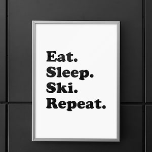 Op de afbeelding: Een ingelijst print met de tekst "Eat. Sleep. Ski. Repeat." in zwarte vetgedrukte letters op een witte achtergrond.