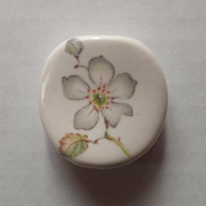 May include: A small, rounded, white ceramic button featuring a hand-painted illustration of a single white flower with five petals, a green and yellow center, a small bud, and a single green leaf on a thin brown stem.