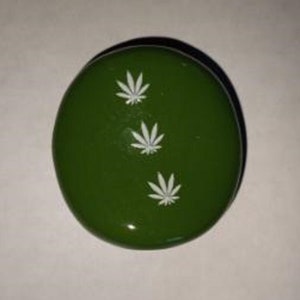 May include: A round, green object with three white cannabis leaf designs. The object has a glossy finish and a dark shadow. The design is simple and the color is a deep, forest green.