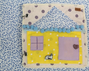 Könnte beinhalten: Ein gelbes und violettes Stoffhaus mit einem blauen Dach und weißen Punkten. Das Haus hat ein Fenster, eine Tür und eine Katze auf der Vorderseite. Das Haus ist an einem weißen Stoff mit blauem Blumendruck befestigt.