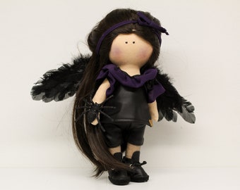 Könnte beinhalten: Eine handgemachte Puppe mit schwarzen Flügeln, einem lila Schal und schwarzer Kleidung. Die Puppe hält eine schwarze Spinne in der Hand.