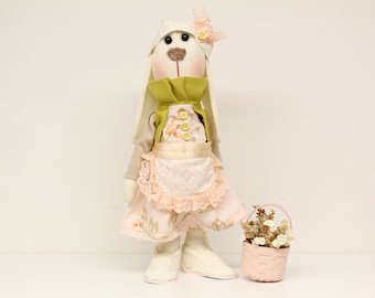 Könnte beinhalten: Eine weiße Stoff-Hasenpuppe, die ein grün-rosa Kleid, einen weißen Hut mit einer rosa Blume und weiße Schuhe trägt. Die Puppe hält einen rosa Korb mit getrockneten Blumen.