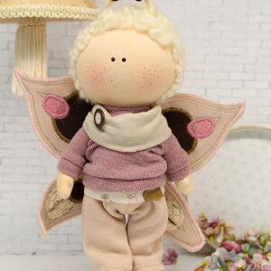 K&ouml;nnte beinhalten: Eine handgemachte Puppe mit einem rosa Pullover, einer beige Hose und Schmetterlingsfl&uuml;geln. Die Puppe hat einen hellbraunen Knopf auf ihrem Pullover und wei&szlig;es Garnhaar.
