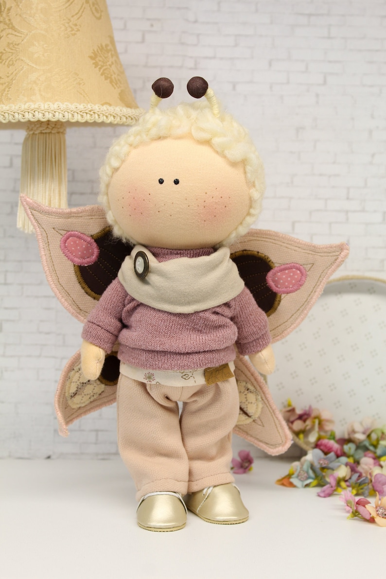 K&ouml;nnte beinhalten: Eine handgemachte Puppe mit einem rosa Pullover, einer beige Hose und Schmetterlingsfl&uuml;geln. Die Puppe hat einen hellbraunen Knopf auf ihrem Pullover und wei&szlig;es Garnhaar.