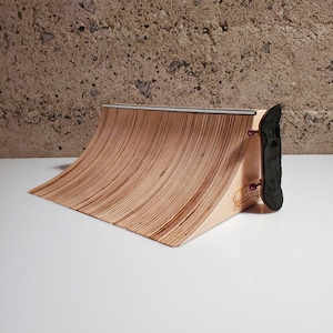 May include: A wooden fingerboard ramp with a black fingerboard on the side. The ramp is made of plywood and has a slight curve. The fingerboard is attached to the ramp with a screw. The ramp is labelled "100% Fingerboard Ready".