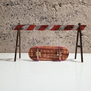 May include: A wooden fingerboard with the words "Dead Blie" printed on it. The fingerboard has purple wheels and is sitting in front of a red and white striped barricade.