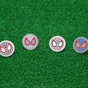 Op de afbeelding: Vier zilveren golfbalmarkers met het Spider-Man maskerontwerp in rood, zwart en blauw. De markers liggen op een groene kunstgras ondergrond.