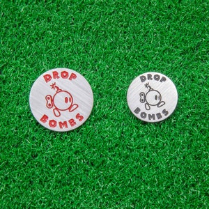 May include: Two silver golf ball markers with the words "DROP BOMBS" and a cartoon bomb graphic. One marker is larger than the other. The markers are on a green surface.