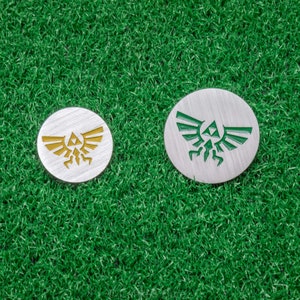 May include: Two silver golf ball markers with a green and gold Triforce symbol. The markers are on a green artificial grass surface.